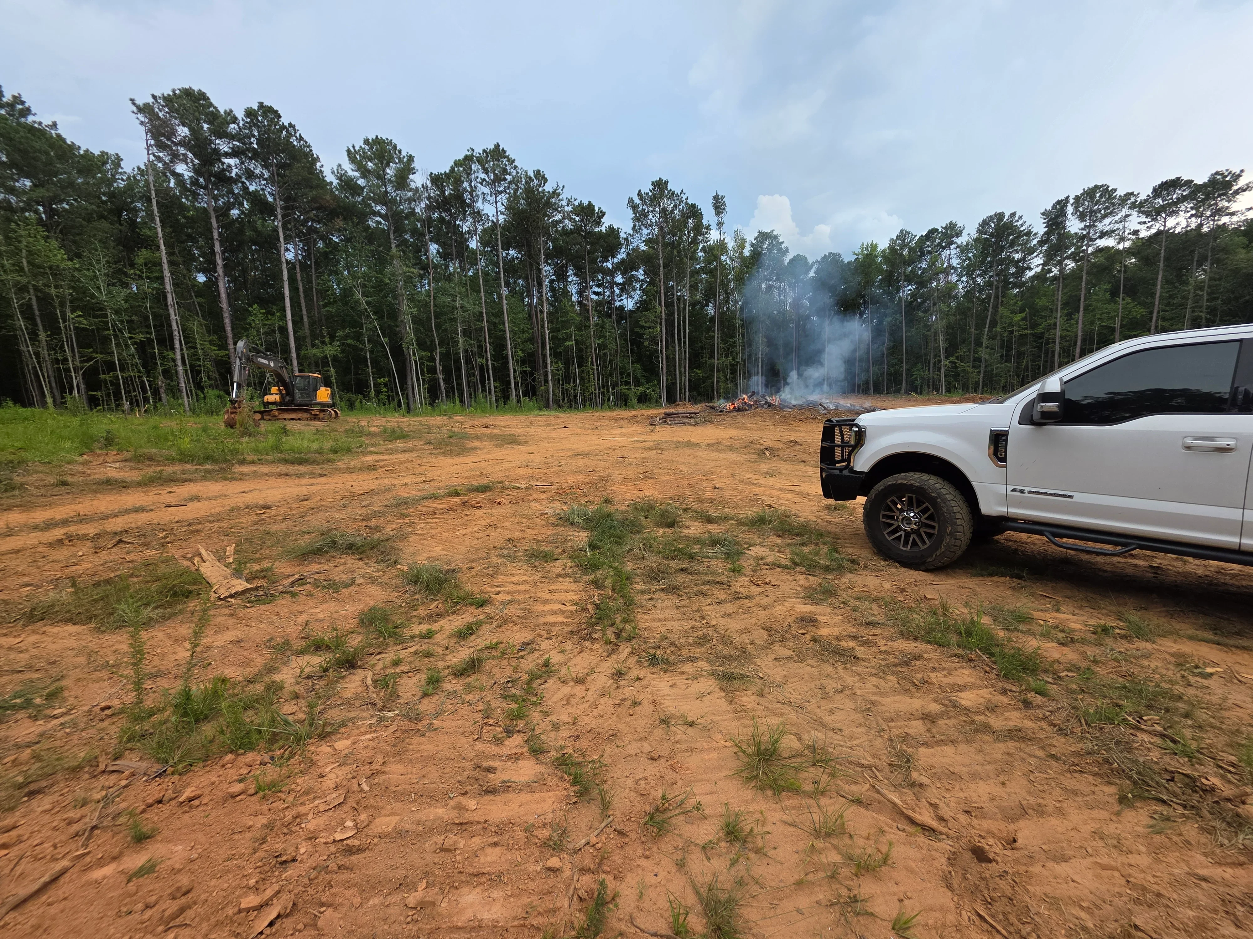 Cleared land with pine trees, heavy equipment, and controlled burn pile during demolition and site prep