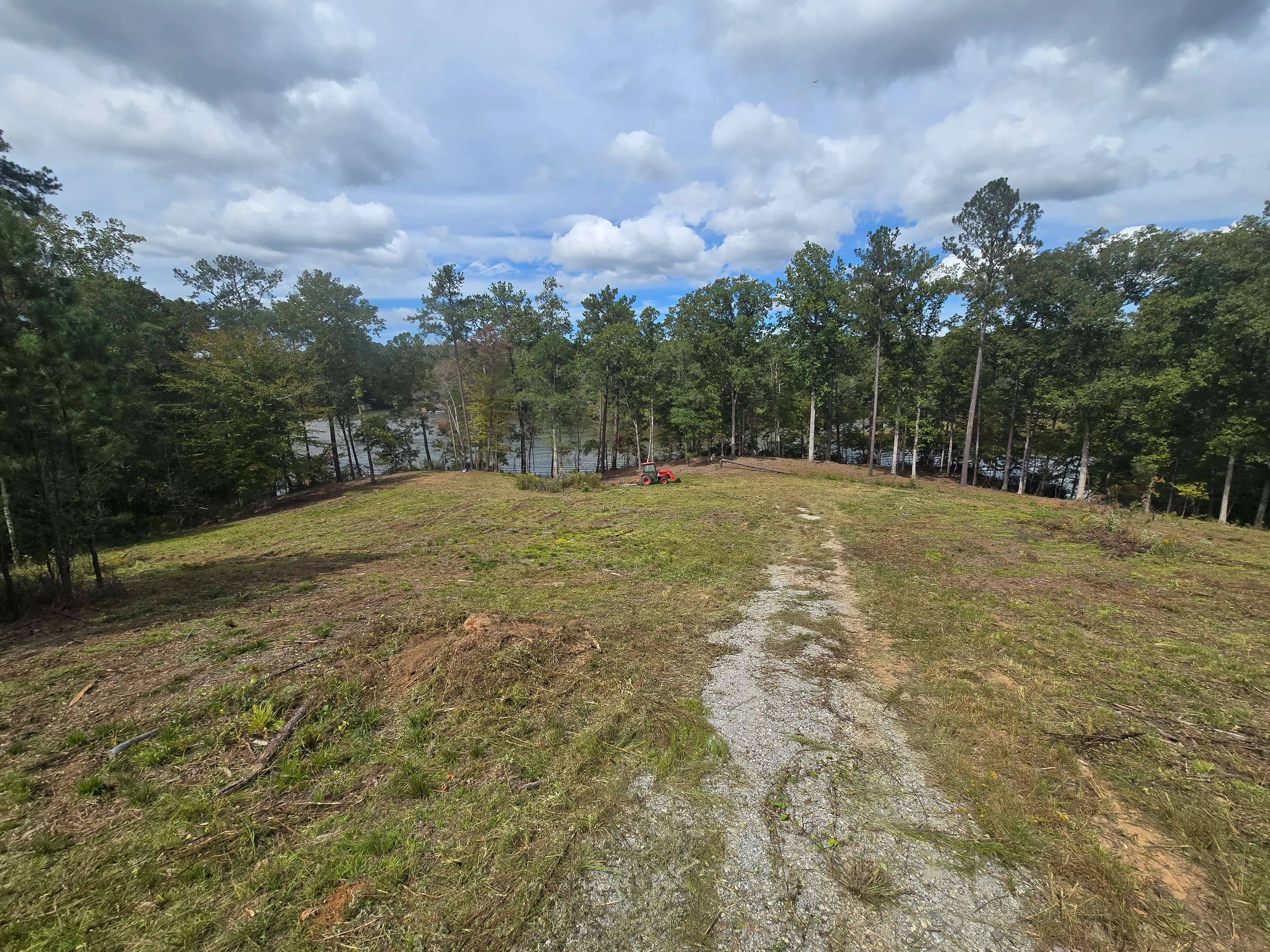 Cleared waterfront lot with gravel road, mature trees retained at edges, and water view in background
