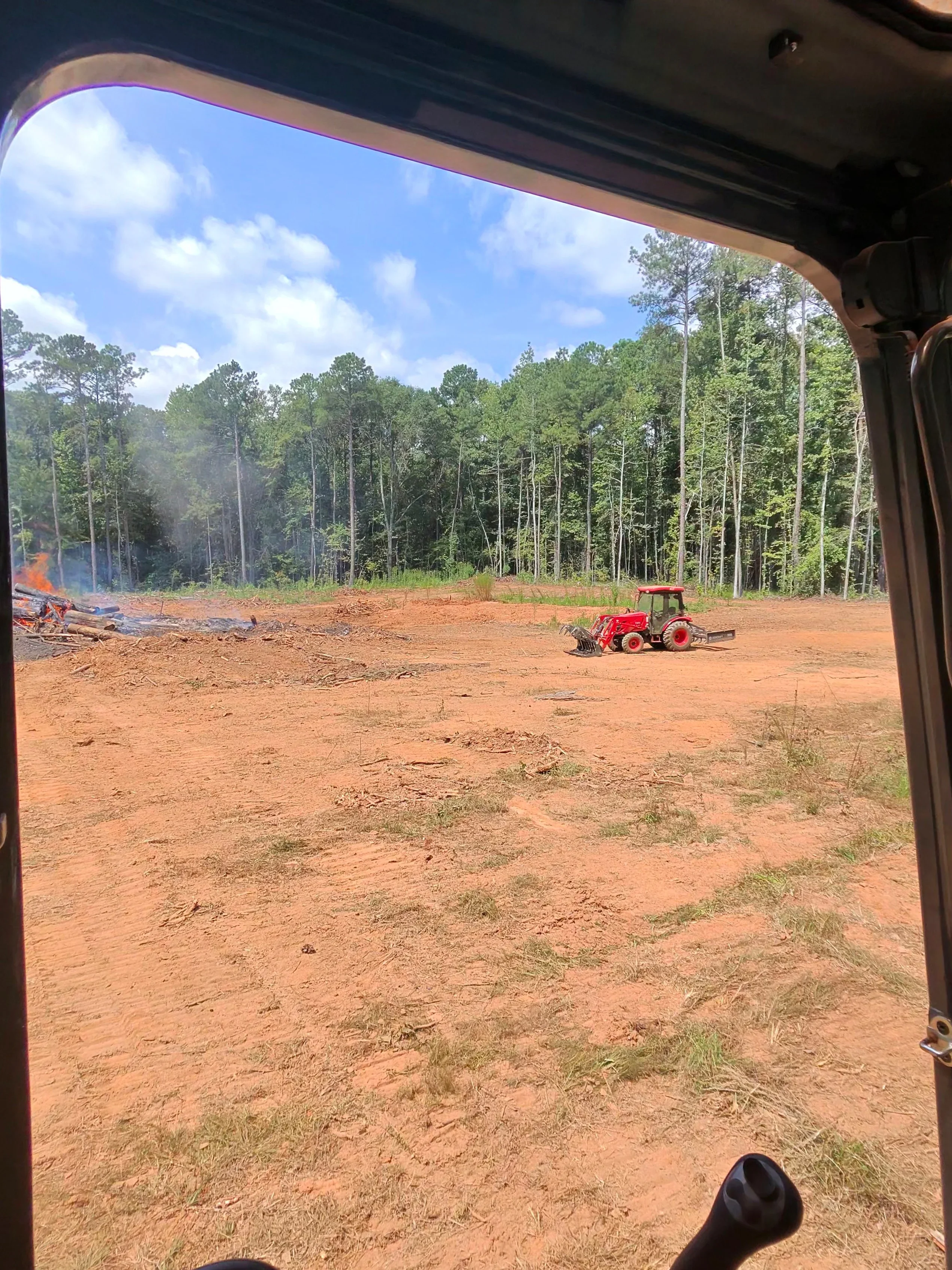 Red grader on cleared dirt lot with timber debris and pine forest in background