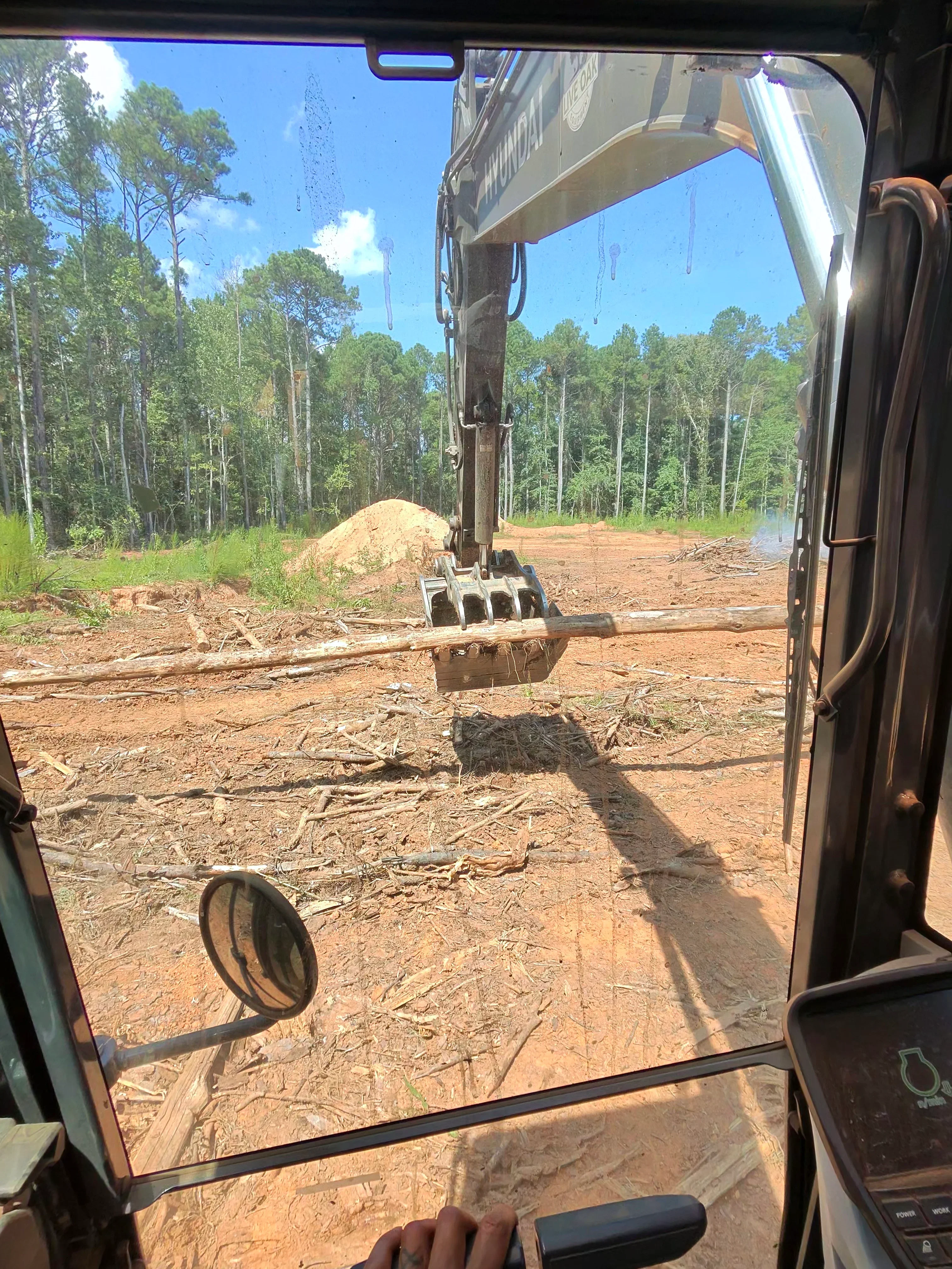 Excavator with four-tine bucket clearing land, removing soil and debris from wooded site