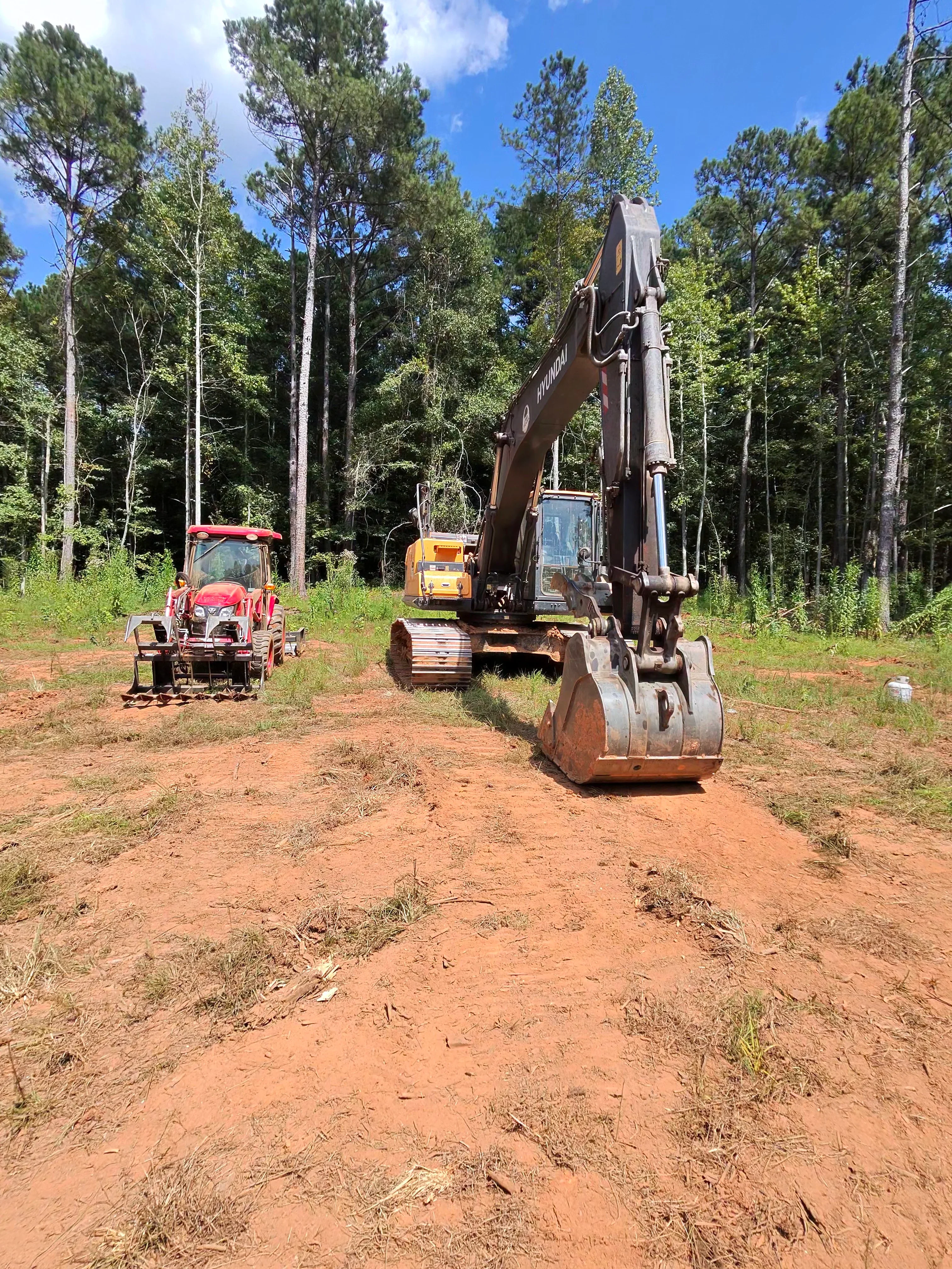 Tracked excavator with compaction drum clearing forested land, red tractor and yellow roller visible on prepared dirt site