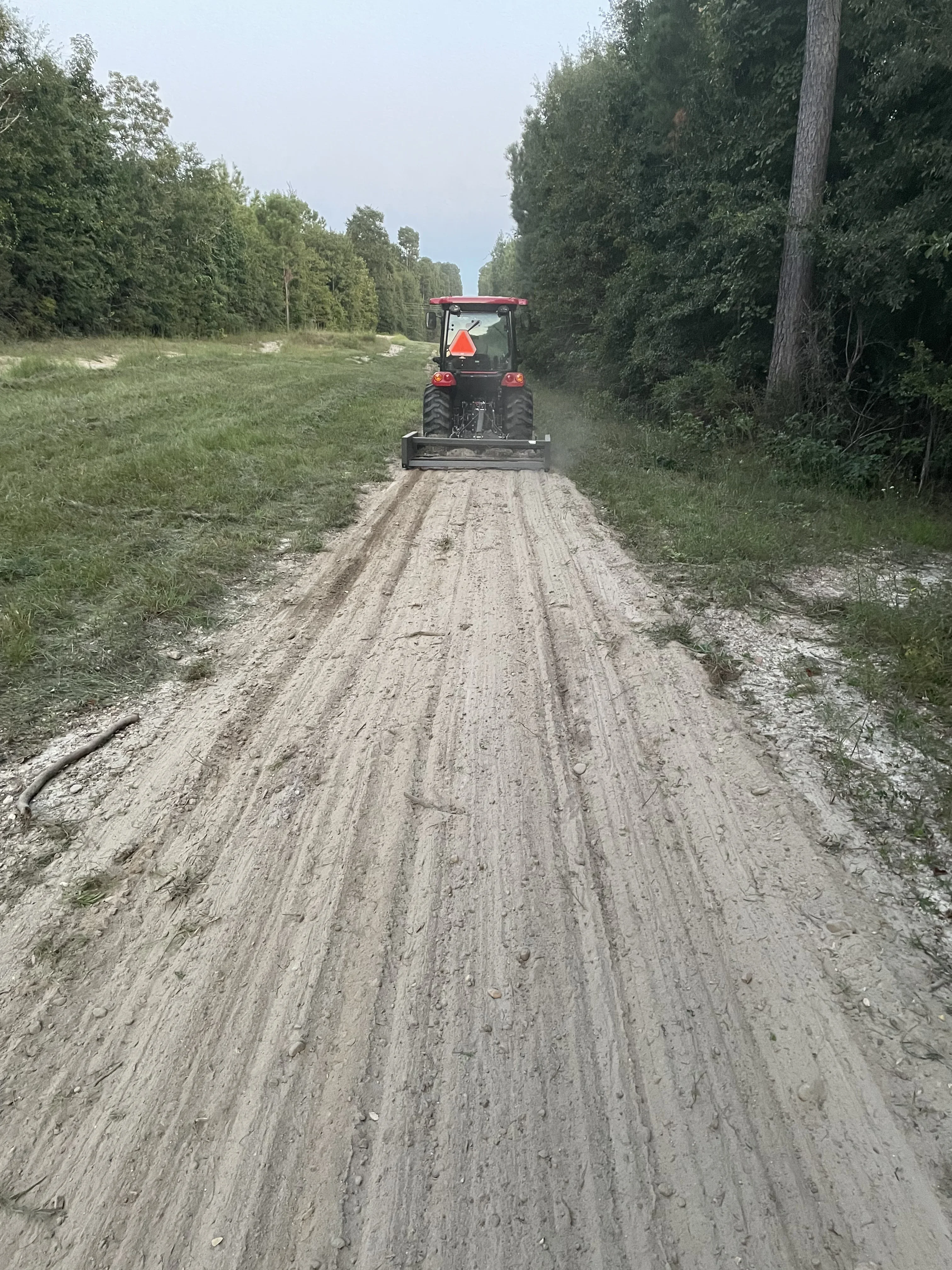 Motor grader compacting gravel road with tree-lined sides
