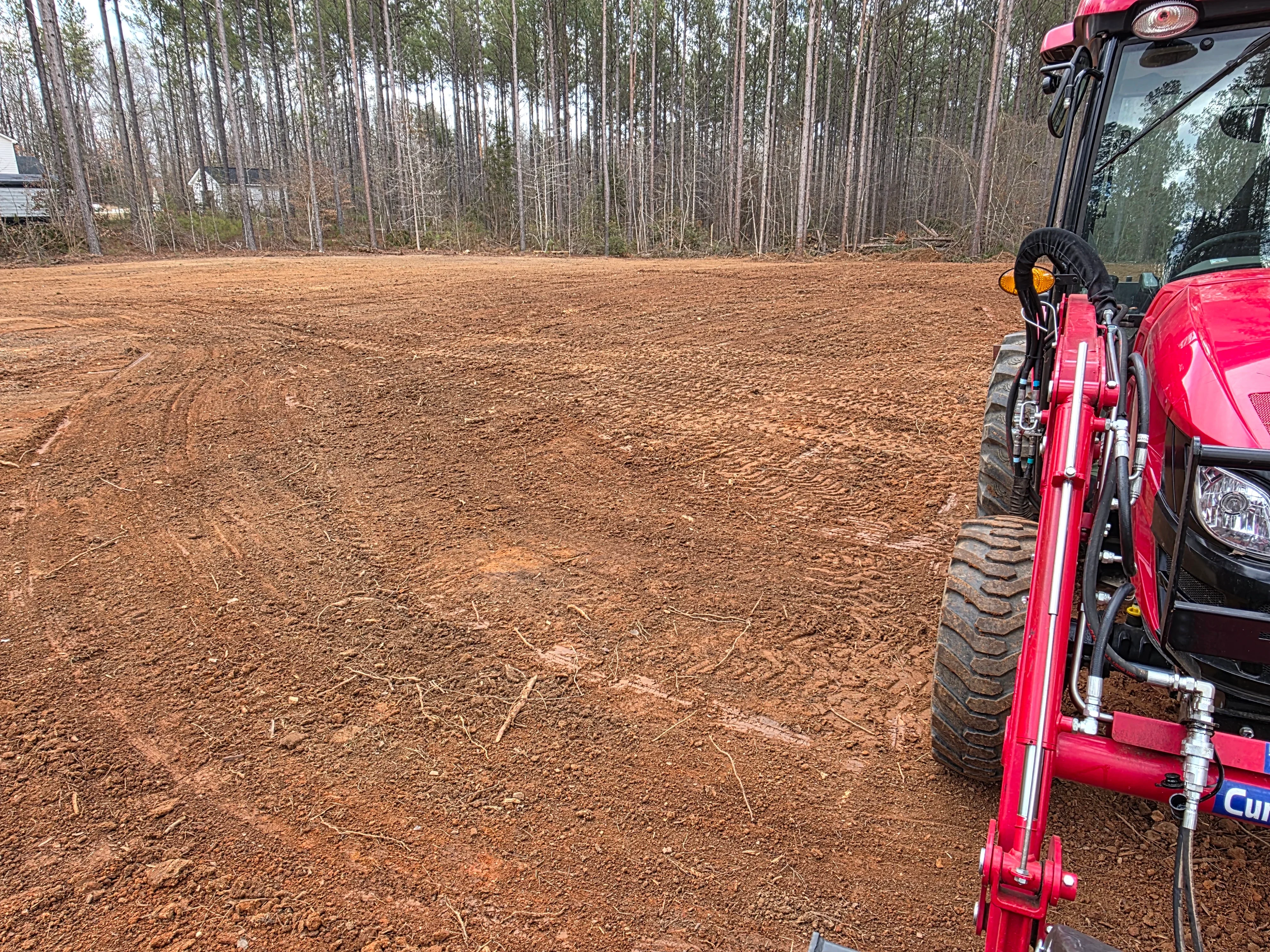 Cleared dirt lot with graded soil and pine forest backdrop, heavy equipment visible