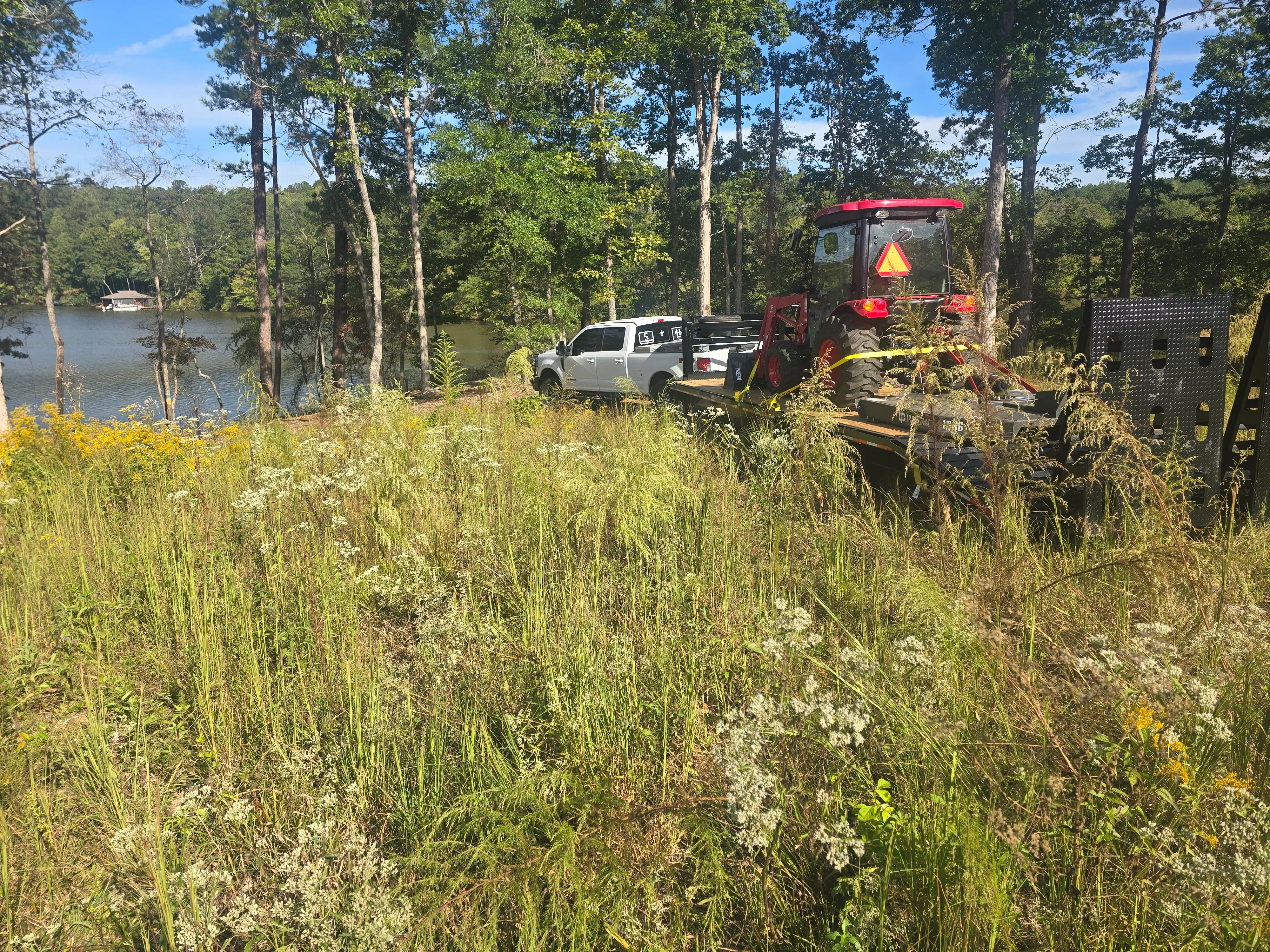 Red excavator clearing overgrown brush and vegetation from waterfront property with support vehicles