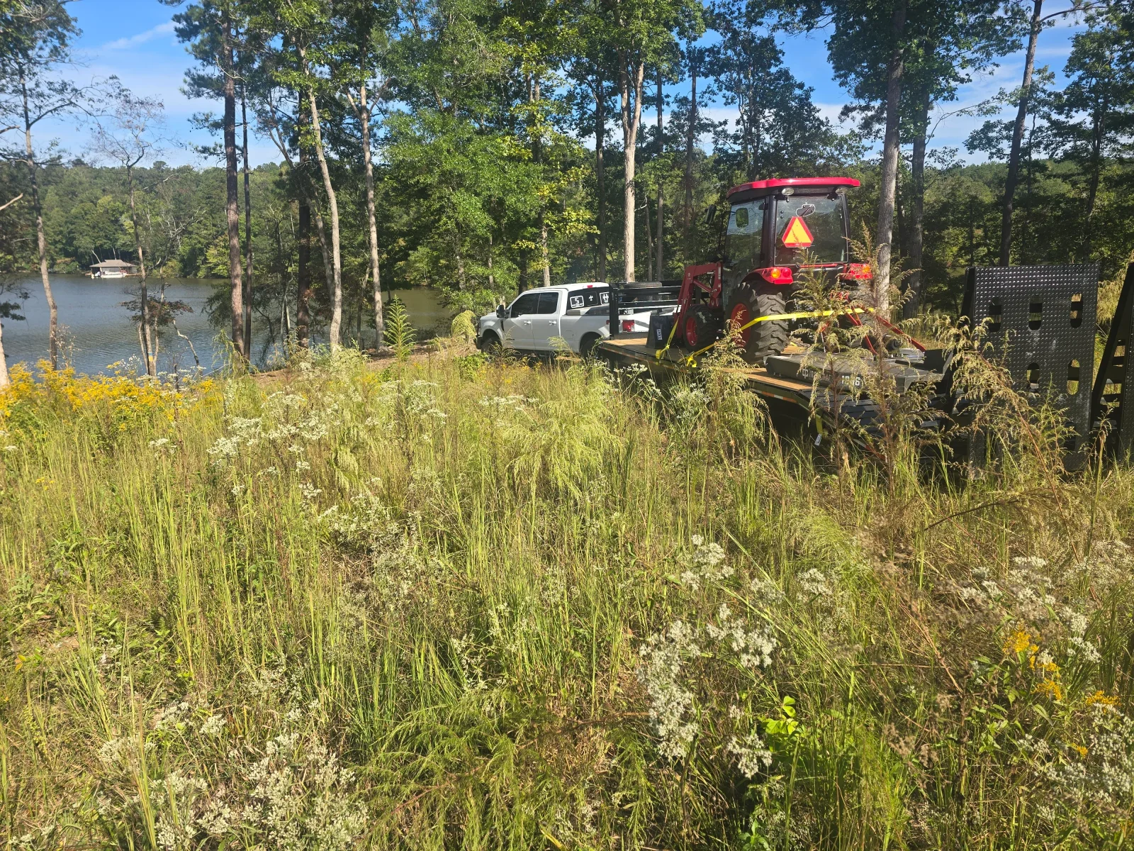 A tractor-mounted rotary cutter clearing a field of tall grass and thick brush on a rural property near Columbus, GA. The cleared area shows flat, open ground where dense overgrowth previously covered the land.